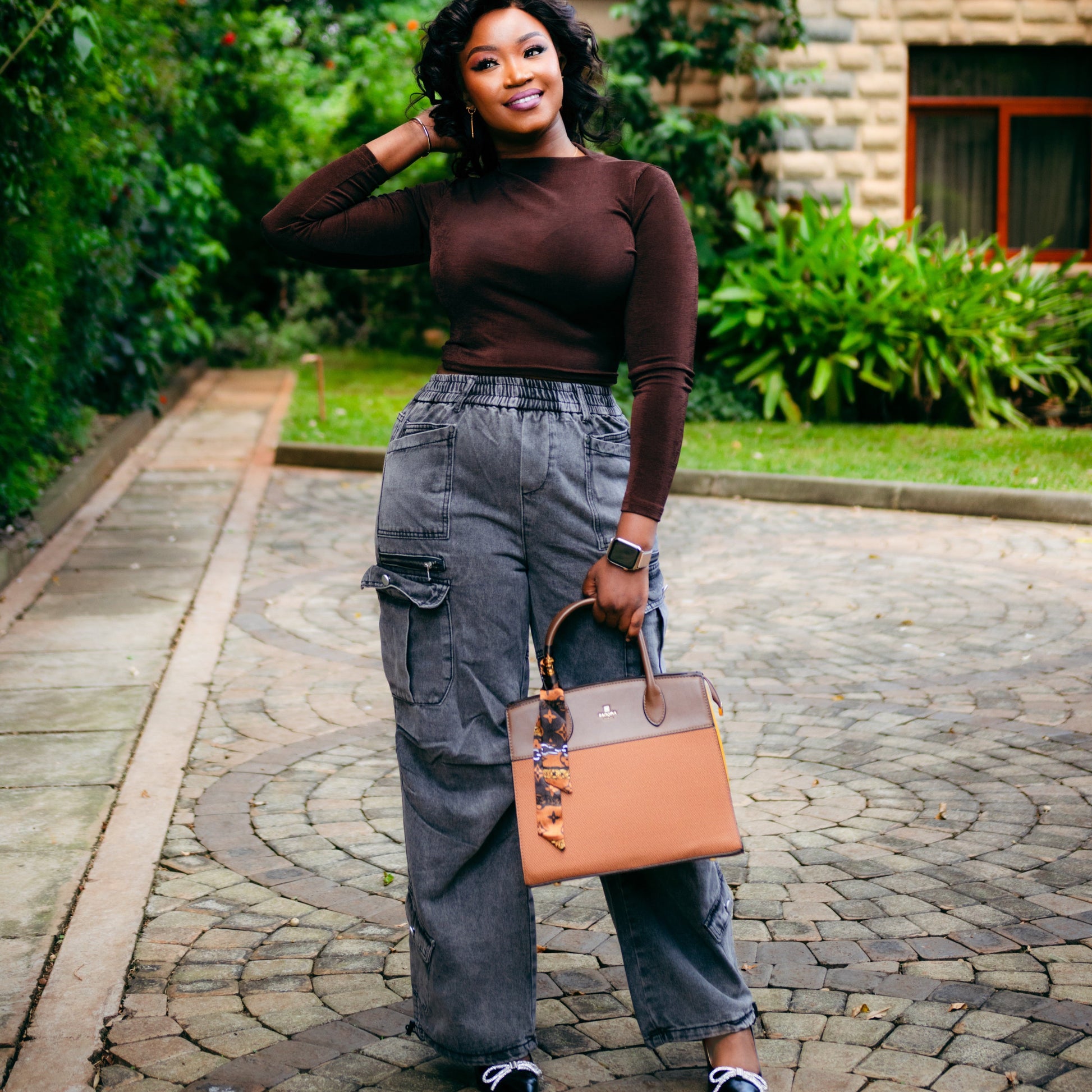 Woman in brown top and gray pants holding a tan handbag outdoors.